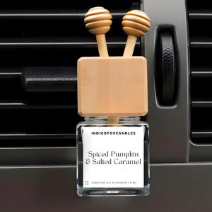 May include: A car air freshener with a glass jar and wooden accents. The label reads "Spiced Pumpkin & Salted Caramel." The diffuser has two wooden toppers with a honey dipper design. The air freshener is displayed against a car vent.
