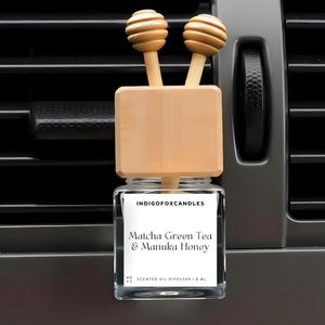 Könnte beinhalten: Ein Auto-Lufterfrischer mit einem Holzaufsatz und zwei dekorativen Holzkugeln. Das Glas hat ein Etikett mit der Aufschrift "Matcha Green Tea & Manuka Honey". Der Lufterfrischer ist für die Platzierung in einer Lüftungsöffnung im Auto konzipiert.