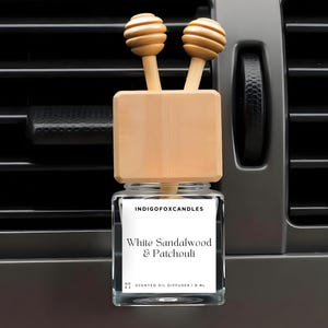 Könnte beinhalten: Ein Auto-Lufterfrischer mit einer transparenten Glasflasche und einem Holzaufsatz. Auf dem Etikett steht "White Sandalwood & Patchouli". Der Holzaufsatz hat zwei dekorative Holzkugeln. Der Lufterfrischer ist für die Befestigung an einer Lüftungsöffnung im Auto konzipiert.