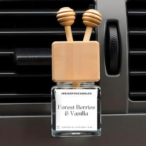 Könnte beinhalten: Ein Auto-Lufterfrischer mit einem Glasbehälter und Holzakzenten. Auf dem Etikett steht "Forest Berries & Vanilla". Der Diffusor hat zwei hölzerne Honiglöffel-Aufsätze. Er ist für die Platzierung in einer Lüftungsöffnung im Auto konzipiert.