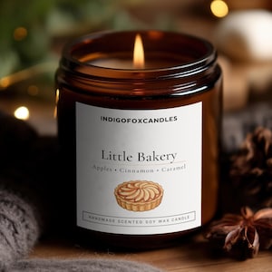 Könnte beinhalten: Eine brennende Sojawachskerze in einem bernsteinfarbenen Glasgefäß mit einem weißen Etikett, das INDIGOFOXCANDLES, Little Bakery, Apples, Cinnamon, Caramel und Handmade Scented Soy Wax Candle sowie eine Illustration einer Apfeltarte zeigt.