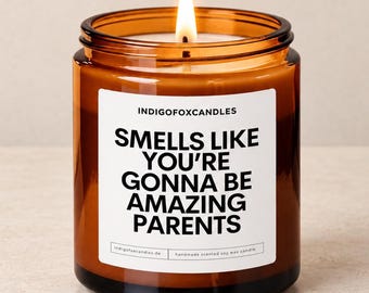 May include: A lit candle in an amber glass jar with a white label featuring the black text INDIGOFOXCANDLES and SMELLS LIKE YOU'RE GONNA BE AMAZING PARENTS. The candle has a single wick and a warm flame, presented in a simple, cylindrical container.