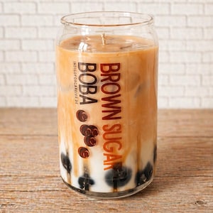 Op de afbeelding: Een glazen kaars met een bruine suiker boba-ontwerp. De kaars heeft een gelaagd uiterlijk, met een witte basis, bruine vloeistof en zwarte boba-parels. De woorden "BROWN SUGAR BOBA" staan op het glas.