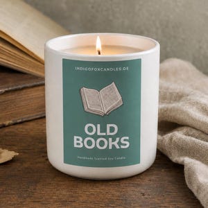 Könnte beinhalten: Eine weiße Kerze im Keramikgefäß mit brennendem Docht. Das Etikett ist blaugrün mit einer schwarzen Strichzeichnung eines offenen Buches und dem Text "INDIGOFOXCANDLES.DE", "OLD BOOKS" und "Handmade Scented Soy Candle" in weißer und schwarzer Schrift.