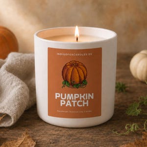 Può includere: Una candela in un vasetto di ceramica bianca con stoppino acceso e un'etichetta rettangolare arancione. L'etichetta presenta un'illustrazione di una zucca e il testo "INDIGOFOXCANDLES.DE", "PUMPKIN PATCH" e "Handmade Scented Soy Candle".