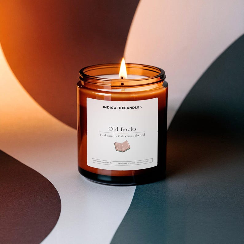 May include: A lit candle in an amber glass jar with a white label reading INDIGOFOXCANDLES, Old Books, Teakwood, Oak, Sandalwood, and featuring a small book icon. The candle has a single wick and is filled with wax, emitting a warm, bright flame.
