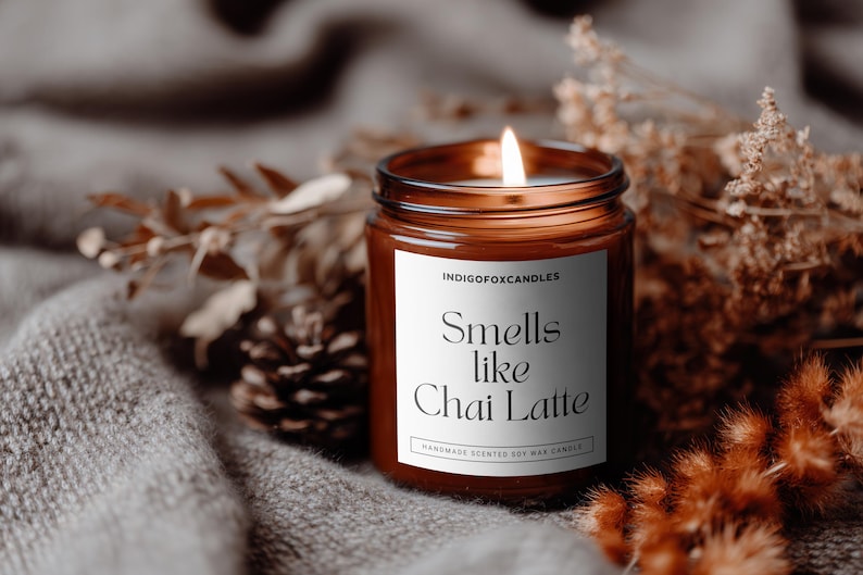Puede incluir: Una vela encendida en un tarro de vidrio marr&oacute;n con una etiqueta blanca que dice "Smells like Chai Latte". La vela est&aacute; rodeada de plantas secas y una pi&ntilde;a sobre una superficie gris texturizada. La vela es una vela de cera de soja perfumada hecha a mano.