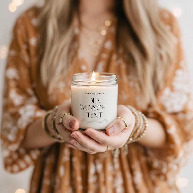 May include: A lit, white candle in a clear glass jar. The candle has the text "DEIN WUNSCH-TEXT" printed on the front. The candle is held in both hands, with gold rings and bracelets visible. The background is blurred.