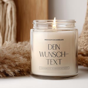 May include: A lit candle in a clear glass jar with the text "DEIN WUNSCH-TEXT" in black. The candle is surrounded by decorative elements, including dried plants and a woven basket. The candle is labeled "INDIGOFoxCANDLES".
