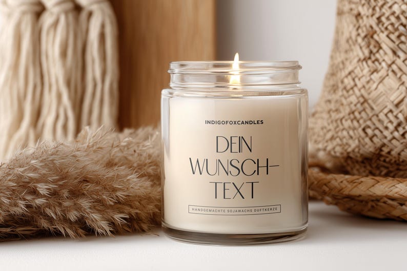 May include: A lit candle in a clear glass jar with the text "DEIN WUNSCH-TEXT" in black. The candle is surrounded by decorative elements, including dried plants and a woven basket. The candle is labeled "INDIGOFoxCANDLES".