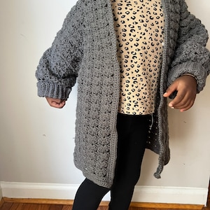 May include: A gray crocheted cardigan with a hood. The cardigan is long and has a textured stitch pattern.
