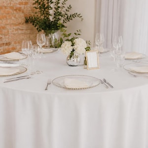May include: A round table set for a formal event, covered with a white tablecloth. Each place setting includes a clear plate with a gold rim, a folded napkin, silverware, and a wine glass. A vase of white flowers and a small framed sign are in the center.