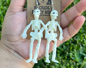 May include: Off-white skeleton earrings with silver hooks. The earrings are held up by a brown tag with the text "PROUDLY PRETTY NINA" and a cartoon illustration. The earrings are made of plastic and are in the shape of a skeleton.