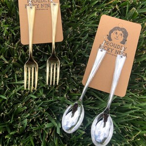 May include: Two pairs of earrings, one gold and one silver, shaped like forks and spoons. The earrings are on brown cards with the text "Proudly picky nina" and a website address.