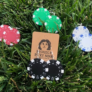 Puede incluir: Un conjunto de cuatro pares de pendientes de fichas de casino. Los pendientes son de color rojo, verde, negro y blanco. Los pendientes están en una tarjeta marrón con el texto "Proudly Pitsky Nina" y una dirección de sitio web.