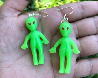 Peut inclure: Une paire de boucles d'oreilles pendantes en forme d'extraterrestre vertes avec des yeux rouges. Les boucles d'oreilles sont en plastique et ont des crochets argentés.