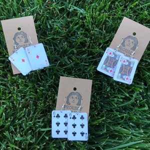 Puede incluir: Tres pares de pendientes con cartas de juego. El primer par es un par de ases de diamantes, el segundo par es un par de reyes de diamantes y el tercer par es un par de seis de tréboles.