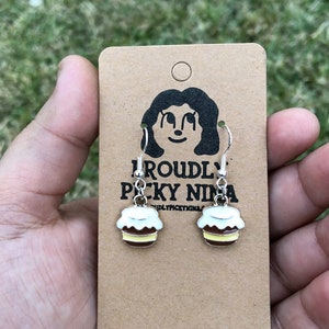 May include: A pair of silver earrings with a yellow, brown, and white cupcake charm. The earrings are on a brown card with the text "Proudly Picky Ninja" printed on it.
