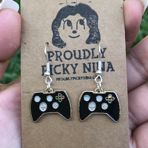 May include: A pair of gold-tone earrings with black video game controller charms. The charms have white and colored buttons. The earrings are on a brown card with the text "Proudly Picky Ninja".