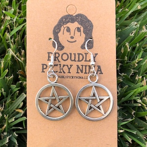 May include: Silver-toned pentagram earrings with a circular frame, displayed on a brown card with the text "PROUDLY PICKY NINA". The earrings hang against a backdrop of green grass.