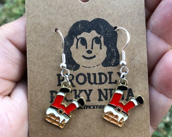 May include: A pair of gold-tone earrings featuring a Santa Claus figure stuck in a chimney. The Santa Claus figure is wearing a red suit with a white trim and a black belt. The earrings are hanging from silver hooks.