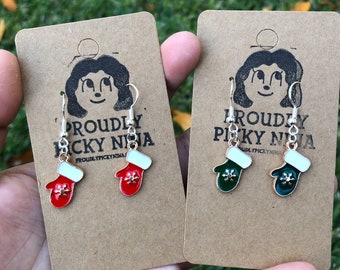 May include: Two pairs of dangle earrings featuring a mitten design. One pair has red mittens with white trim and a snowflake design. The other pair has green mittens with white trim and a snowflake design. The earrings are on a brown card with the text "Proudly Picky Ninja" printed on it.