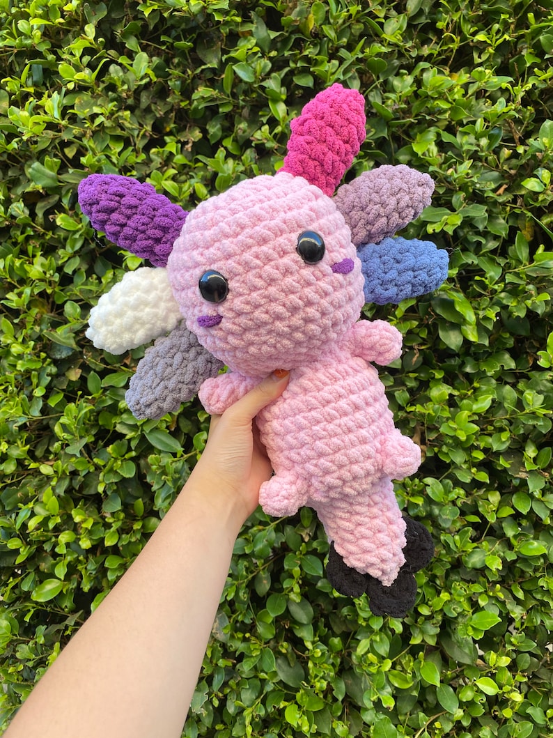 May include: A crocheted axolotl plush toy with a pink body and multi-colored fins. The fins are purple, white, gray, and pink.