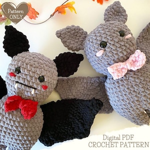 Könnte beinhalten: Zwei gehäkelte Fledermaus-Amigurumi-Spielzeuge. Eine Fledermaus ist grau mit schwarzen Flügeln und einer roten Schleife. Die andere Fledermaus ist grau mit einer rosa Schleife. Die Fledermäuse befinden sich auf einem weißen Hintergrund mit Herbstblättern.