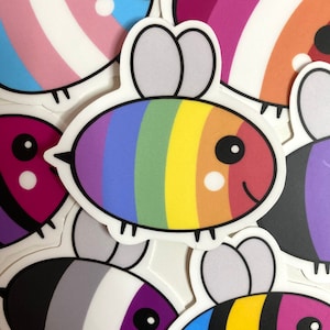 Puede incluir: Un conjunto de pegatinas de abejas coloridas con diferentes diseños de banderas del orgullo. Las pegatinas representan una abeja de dibujos animados con un cuerpo rayado y alas. Los diseños incluyen las banderas del orgullo transgénero, lesbiana, gay, bisexual, asexual y pansexual.