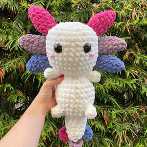 May include: A white crocheted axolotl plush toy with pink, purple, and blue crocheted accents. The toy has large black eyes and a small pink nose.