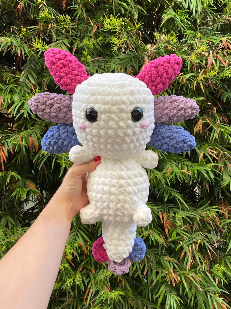 May include: A white crocheted axolotl plush toy with pink, purple, and blue crocheted accents. The toy has large black eyes and a small pink nose.