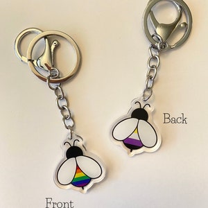 May include: A keychain with a bee design. The front of the bee has a rainbow stripe, and the back has a purple, white, and black stripe. The keychain is attached to a silver chain and a silver ring.
