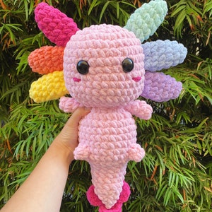 May include: A pink crocheted axolotl plush toy with rainbow-colored crocheted gills. The axolotl has large black eyes and a small pink mouth.