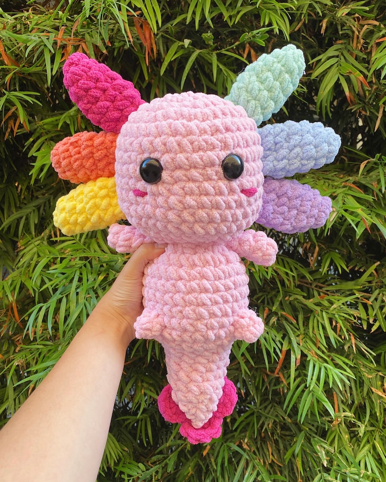 May include: A pink crocheted axolotl plush toy with rainbow-colored crocheted gills. The axolotl has large black eyes and a small pink mouth.
