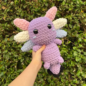 May include: A crocheted axolotl plush toy with purple, pink, white, and blue yarn. The toy has large black eyes and a small mouth.