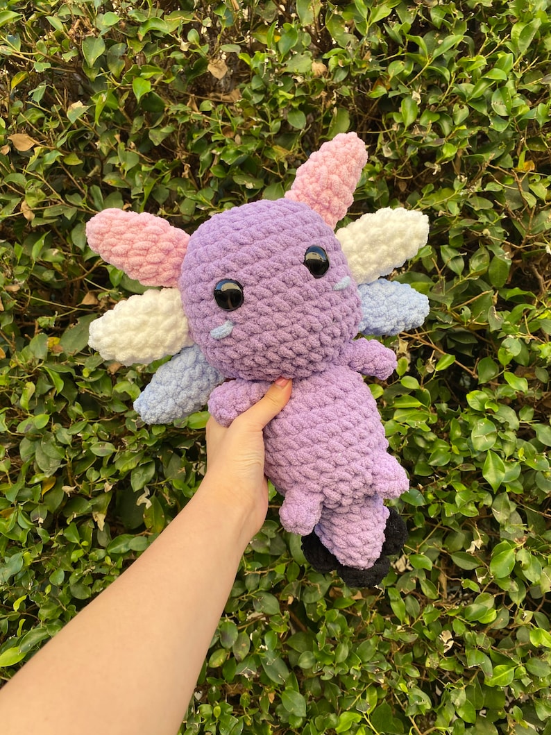 May include: A crocheted axolotl plush toy with purple, pink, white, and blue yarn. The toy has large black eyes and a small mouth.