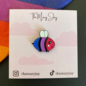 Op de afbeelding: Een bi pride-bij-emaillepin met een blauw, paars en roze gestreept lichaam, witte vleugels en een rood gezicht met witte stippen. De pin is op een roze achtergrond met witte wolken en de tekst "The Mary Jay" bovenaan.