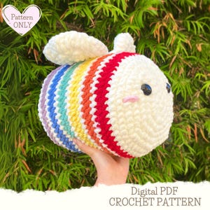 Op de afbeelding: Een haakpatroon voor een regenboog-bij amigurumi. Het patroon is beschikbaar als digitale PDF-download. De bij is wit met een regenboogstrepenpatroon en heeft een schattig gezicht.