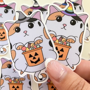 May include: A set of Halloween cat stickers with a calico cat wearing a witch hat and holding a pumpkin bucket filled with candy. The cat has a rainbow hat and is holding a pumpkin bucket filled with candy.