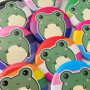 Op de afbeelding: Een verzameling kleurrijke ronde knopen met cartoon-kikkers met verschillende regenboogvlag-ontwerpen. De knopen zijn ongeveer 2,5 cm in diameter en hebben een glanzende afwerking.