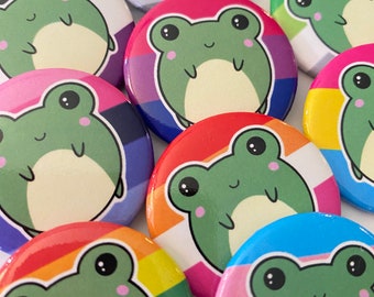 May include: A collection of colorful round buttons featuring cartoon frogs with different pride flag designs. The buttons are approximately 1 inch in diameter and have a shiny finish.