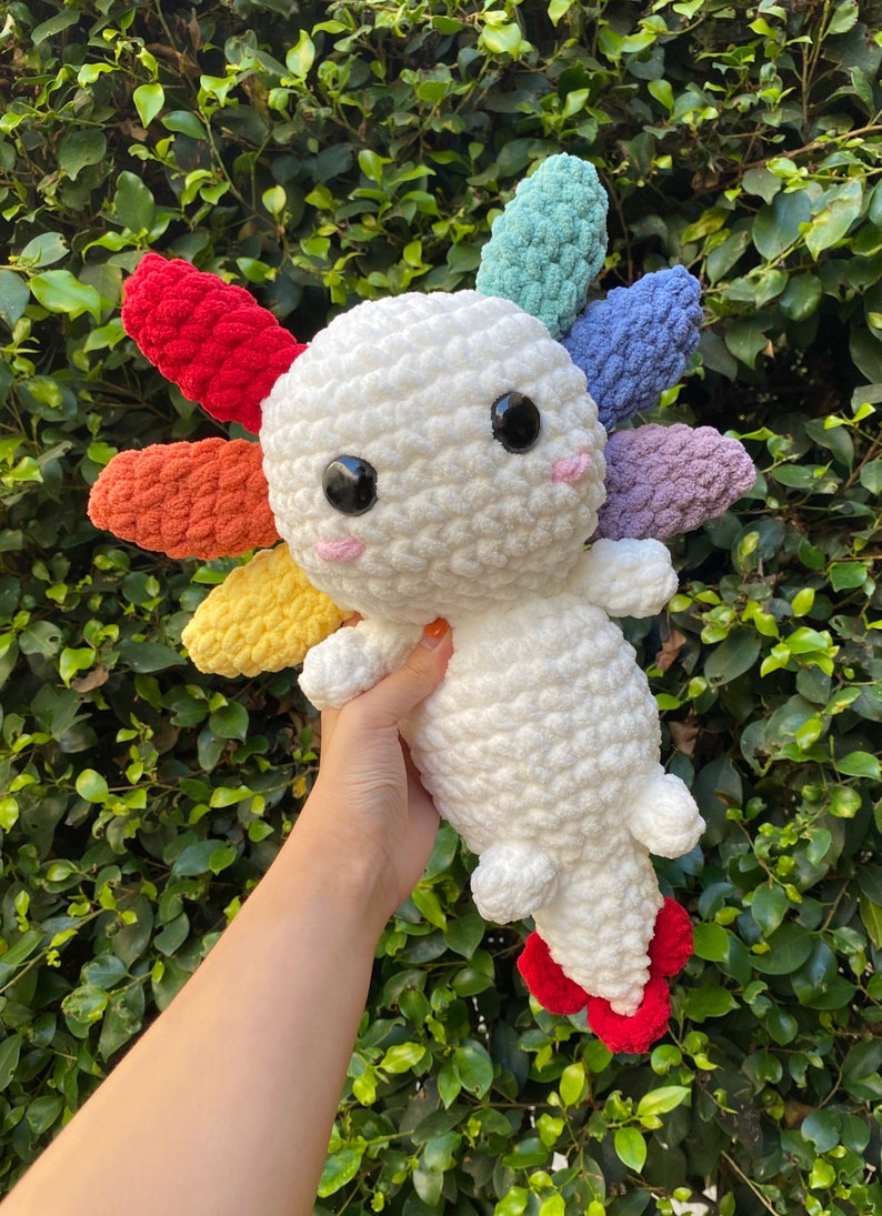 May include: A white crocheted axolotl plush toy with rainbow colored crocheted gills. The axolotl is holding its tail up and has black eyes and a pink nose.