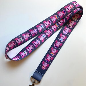 Könnte beinhalten: Ein dunkelgraues Lanyard mit einem sich wiederholenden Muster aus rosa Axolotl, die Pride-Flaggen tragen. Das Lanyard hat einen silbernen Metallclip.