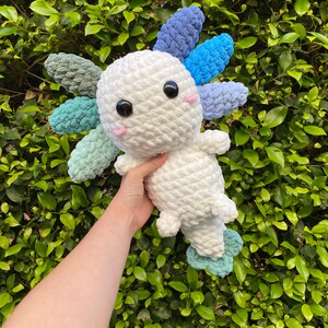 May include: A white crocheted axolotl plush toy with blue, green, and purple crocheted gills. The toy is being held in a hand against a green leafy background.