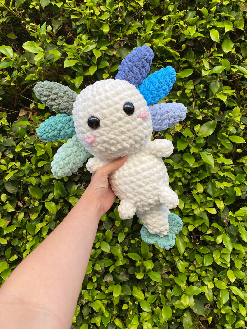 May include: A white crocheted axolotl plush toy with blue, green, and purple crocheted gills. The toy is being held in a hand against a green leafy background.