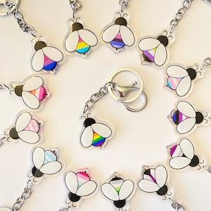 May include: A collection of 13 bee-shaped keychains with different pride flag designs. Each keychain features a clear acrylic bee with a colorful flag design on its abdomen. The keychains are attached to silver metal chains with a lobster clasp.
