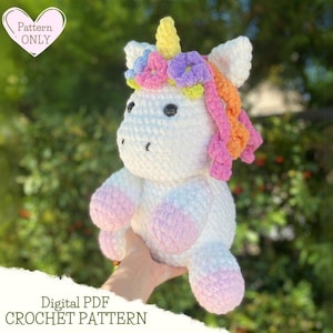 Può includere: Un giocattolo amigurumi a forma di unicorno all'uncinetto con un corpo bianco, zoccoli rosa e una criniera e un corno arcobaleno. L'unicorno indossa una corona di fiori con fiori rosa, viola e gialli.
