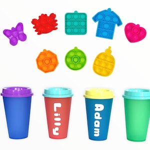 May include: A collection of colorful pop-it toys in various shapes, including a butterfly, heart, and shirt. Below are four reusable coffee cups in blue, red, yellow, and green, with names printed on two of them.