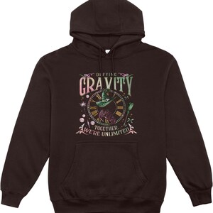 May include: A dark brown hooded sweatshirt featuring a graphic with the words "Defying Gravity" and "Together, We're Unlimited." The graphic includes a witch on a broom, a clock, and decorative elements in shades of green, pink, and gold.