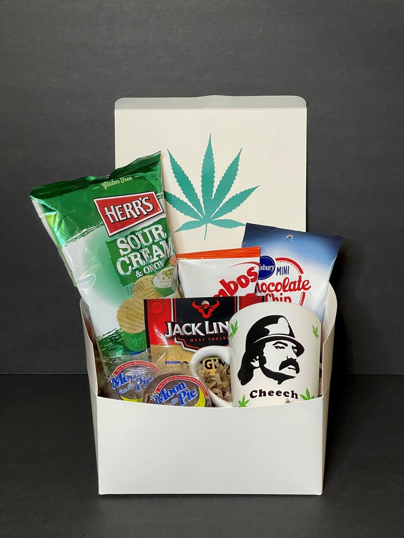 May include: A white gift box filled with snacks and a mug. The box has a green cannabis leaf design. Snacks include chips, cookies, and meat snacks. The mug features a black and white image of a man with the name "Cheech".